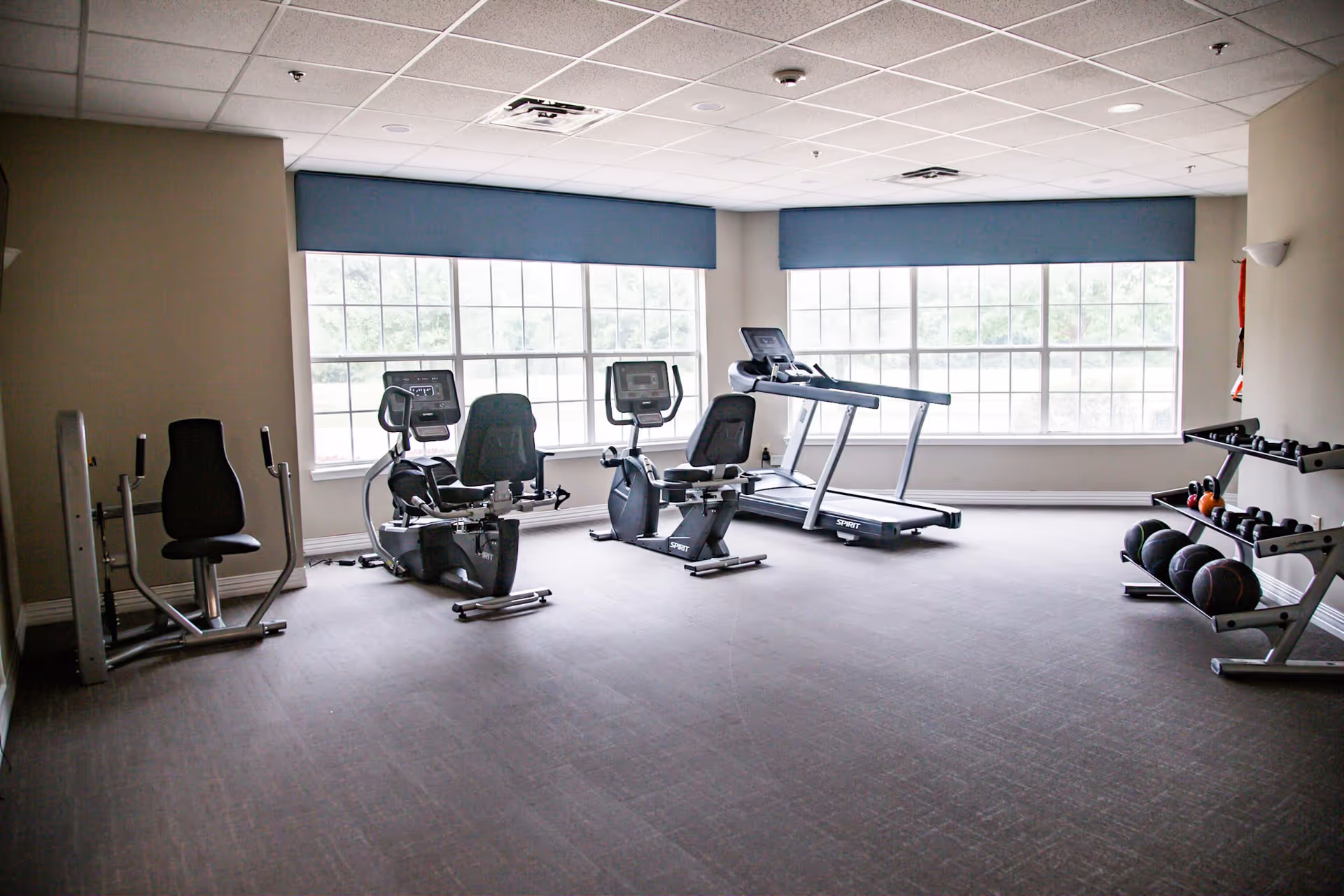 A fitness room with exercise equipment including two recumbent stationary bikes, a treadmill, a weight machine, and racks holding medicine balls and dumbbells. Large windows with blue valances allow natural light into the room.