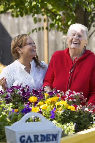 Two women smiling and enjoying time together outdoors near a garden bed filled with colorful flowers, with a white container labeled 'GARDEN' in the foreground.