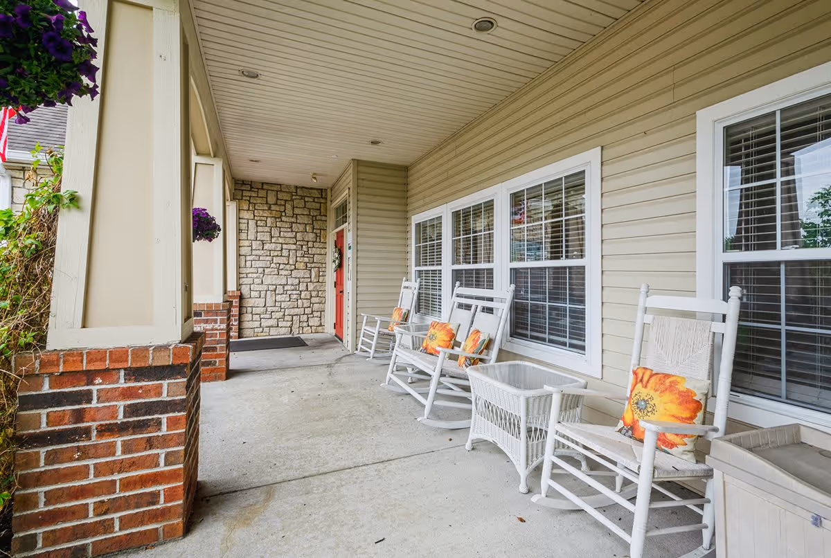A covered outdoor porch area with white rocking chairs adorned with orange floral cushions, a small white wicker table, and beige siding walls with multiple windows. The porch has a concrete floor, brick and stone accents, and a red front door at the end.