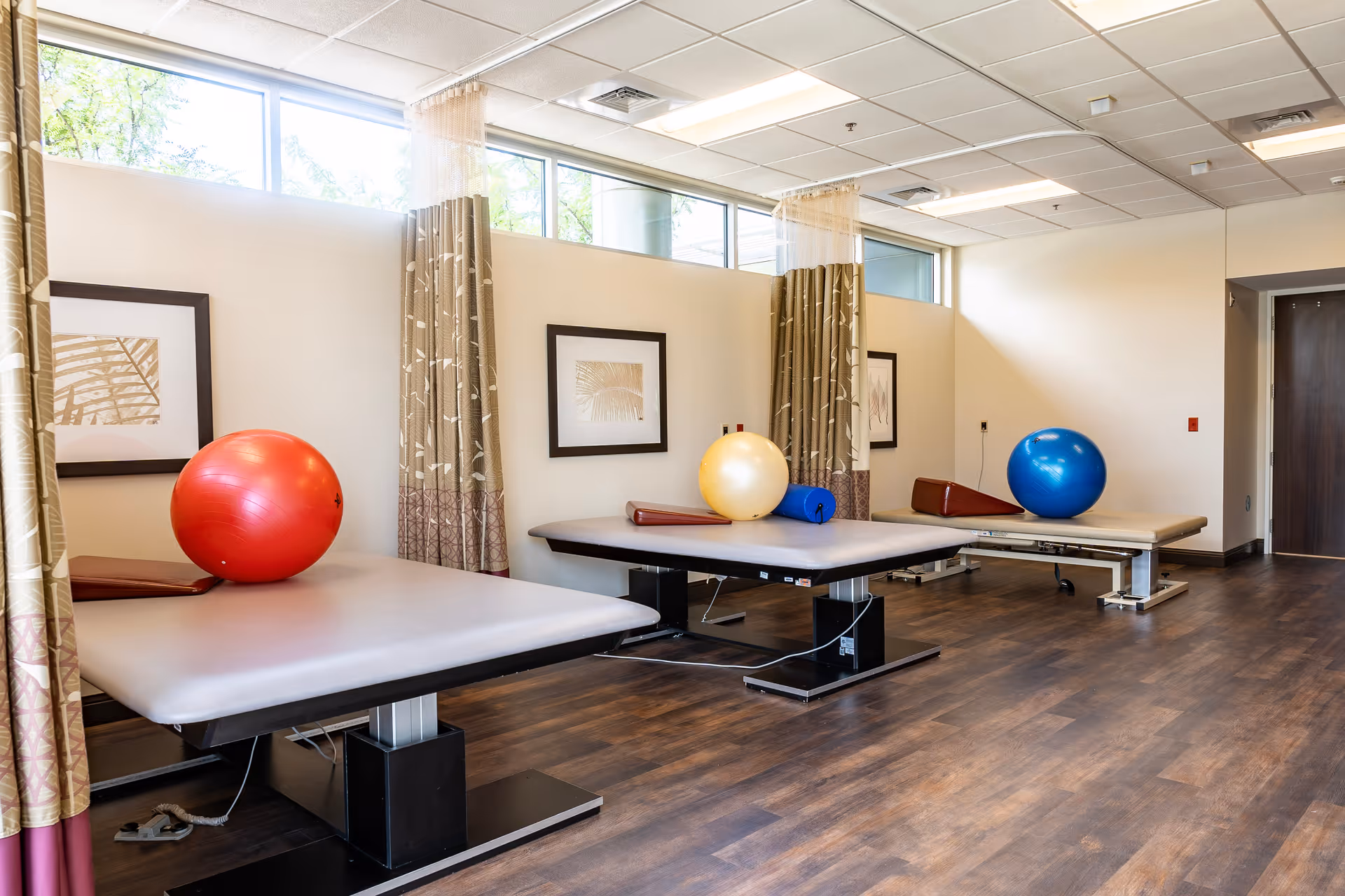 A physical therapy room with three padded treatment tables, each equipped with an exercise ball and a bolster. The room has large windows near the ceiling allowing natural light, framed artwork on the walls, and curtains between the tables for privacy. The floor is wooden, and the overall atmosphere is clean and well-lit.