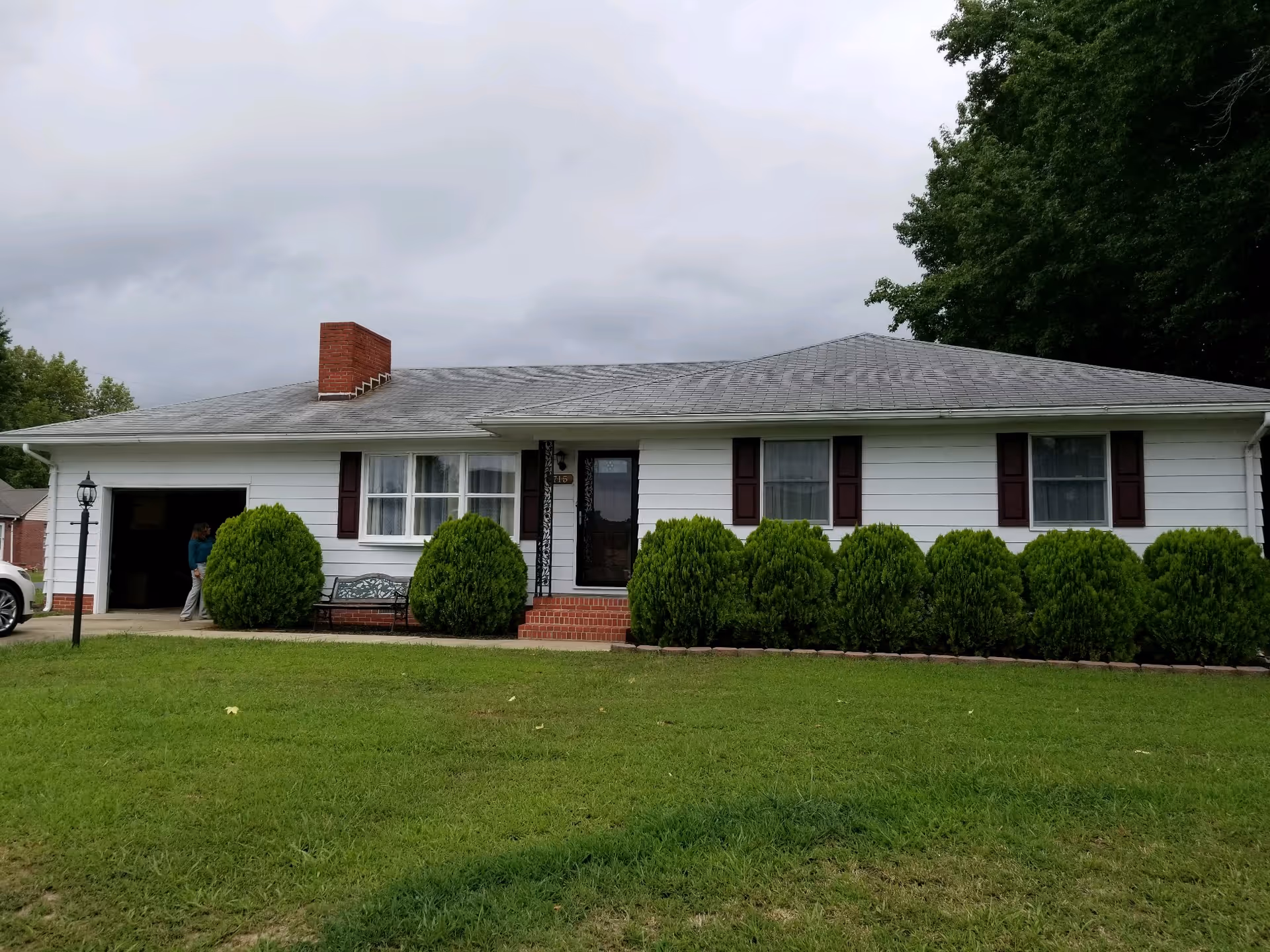 Single-story white house with a gray roof, a brick chimney, and a front porch with steps. There are several green bushes lining the front of the house and a green lawn in the foreground. A person is standing near the open garage on the left side of the house. The sky is overcast.