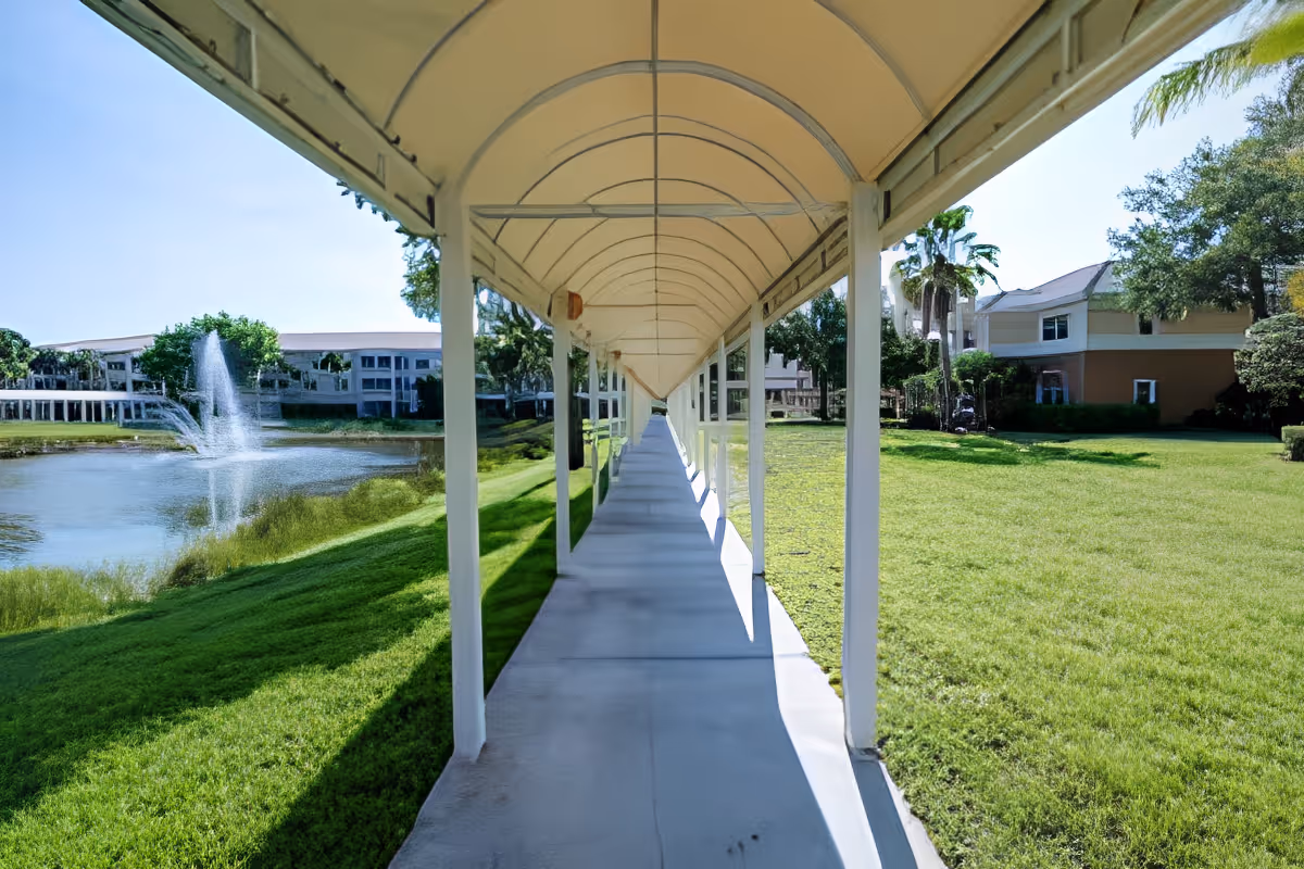 Covered walkway with a white canopy extending over a concrete path, surrounded by green grass and trees. To the left, there is a pond with a water fountain, and buildings are visible in the background under a clear blue sky.