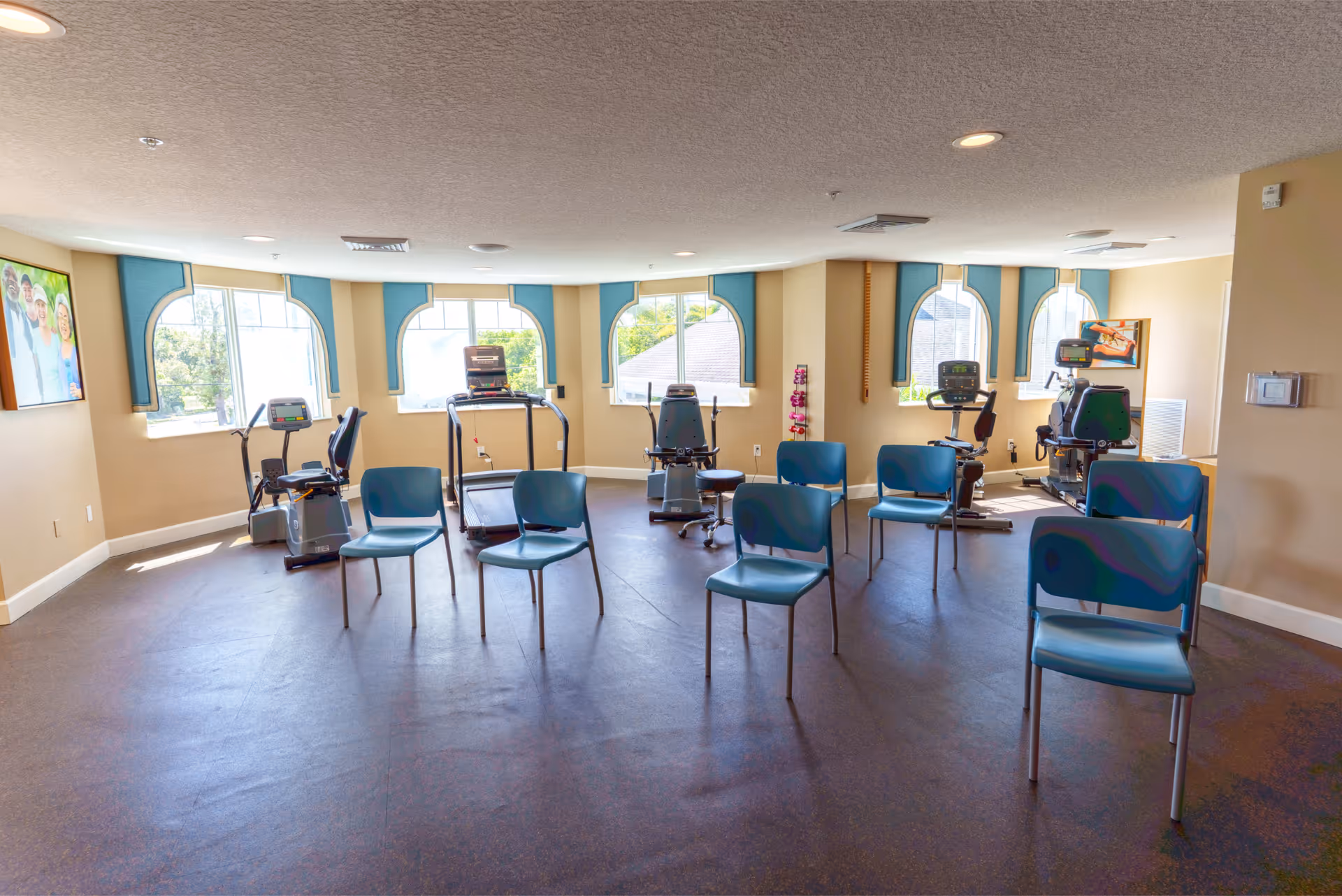 Bright interior exercise room with stationary bikes, treadmills and several blue chairs arranged facing the windows.
