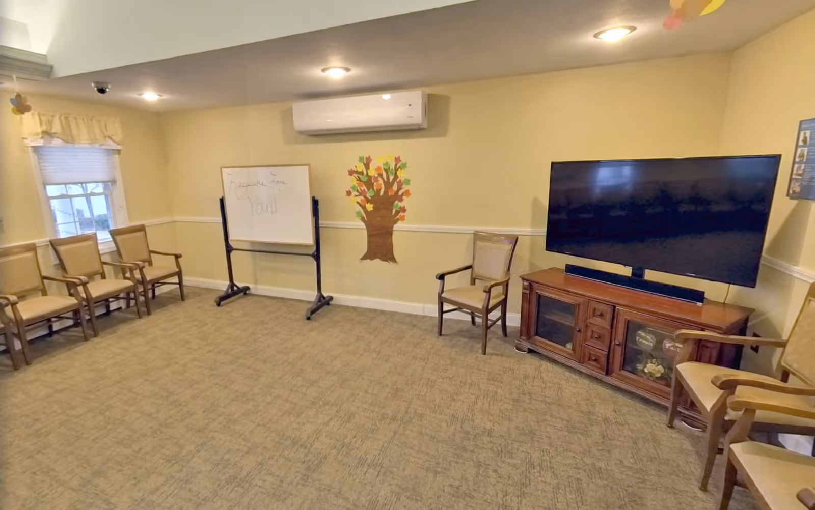 A small meeting or activity room with beige walls and carpet. There are several wooden chairs with beige cushions arranged along the walls. A large flat-screen TV sits on a wooden cabinet on the right side. A whiteboard on a stand is positioned near the center back wall, with a colorful paper tree decoration next to it. A window with a valance is on the left wall, and a ceiling-mounted air conditioning unit is above the whiteboard.