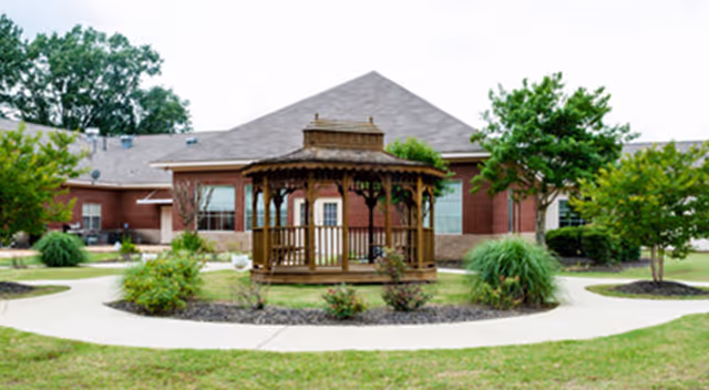 Outdoor view of a senior living facility with a wooden gazebo in the center surrounded by a circular concrete walkway, green grass, and various bushes and trees. The building in the background is made of red brick with large windows and a gray roof.