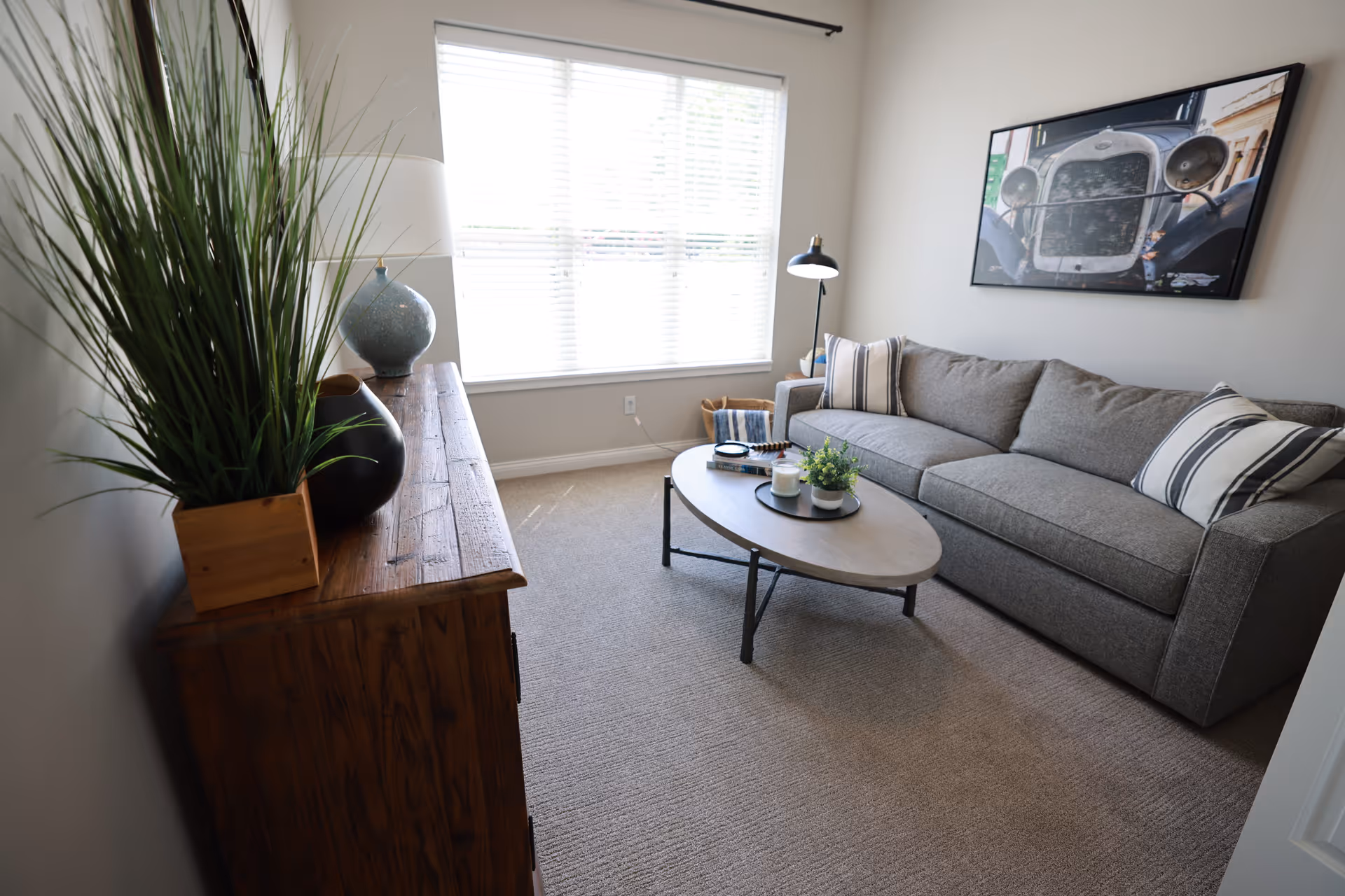 A bright living room with a gray sofa, oval coffee table, wooden console with plants, and a large window.