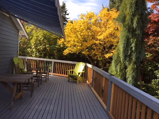 A wooden outdoor deck with a railing, featuring several chairs and a small table. The deck is attached to a building with gray siding and a blue awning. Surrounding the deck are trees with green, yellow, and red autumn foliage under a partly cloudy sky.