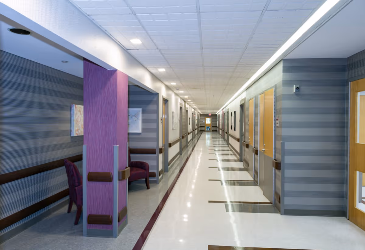Long, well-lit interior hallway of a healthcare facility with seating alcoves, patterned walls and doors lining the corridor.