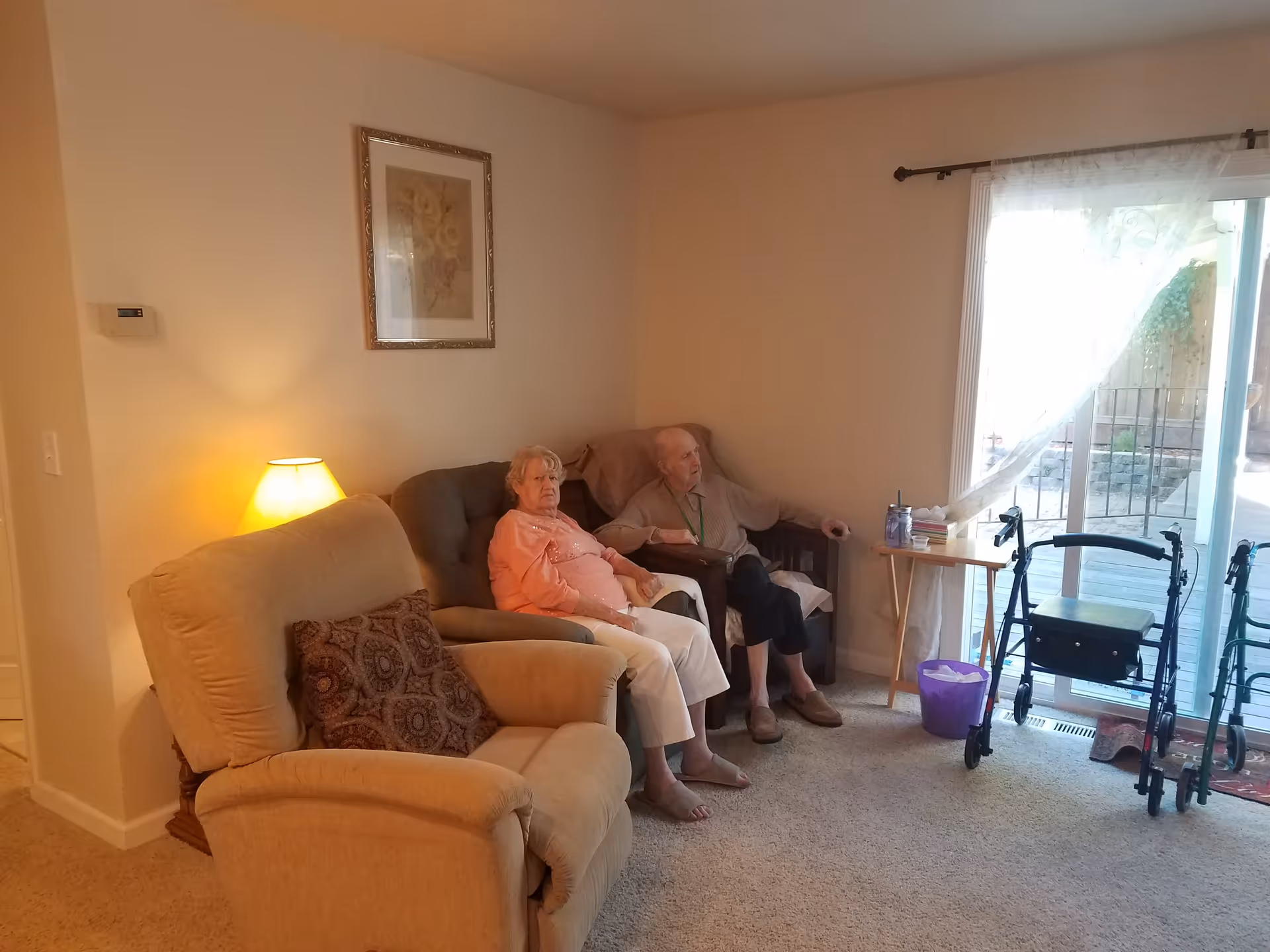 A living room with two elderly people sitting on chairs. There is a beige recliner with a patterned pillow, a lamp on a side table, a framed picture on the wall, and a sliding glass door with sheer curtains leading to an outdoor area. Two walkers are positioned near the door.