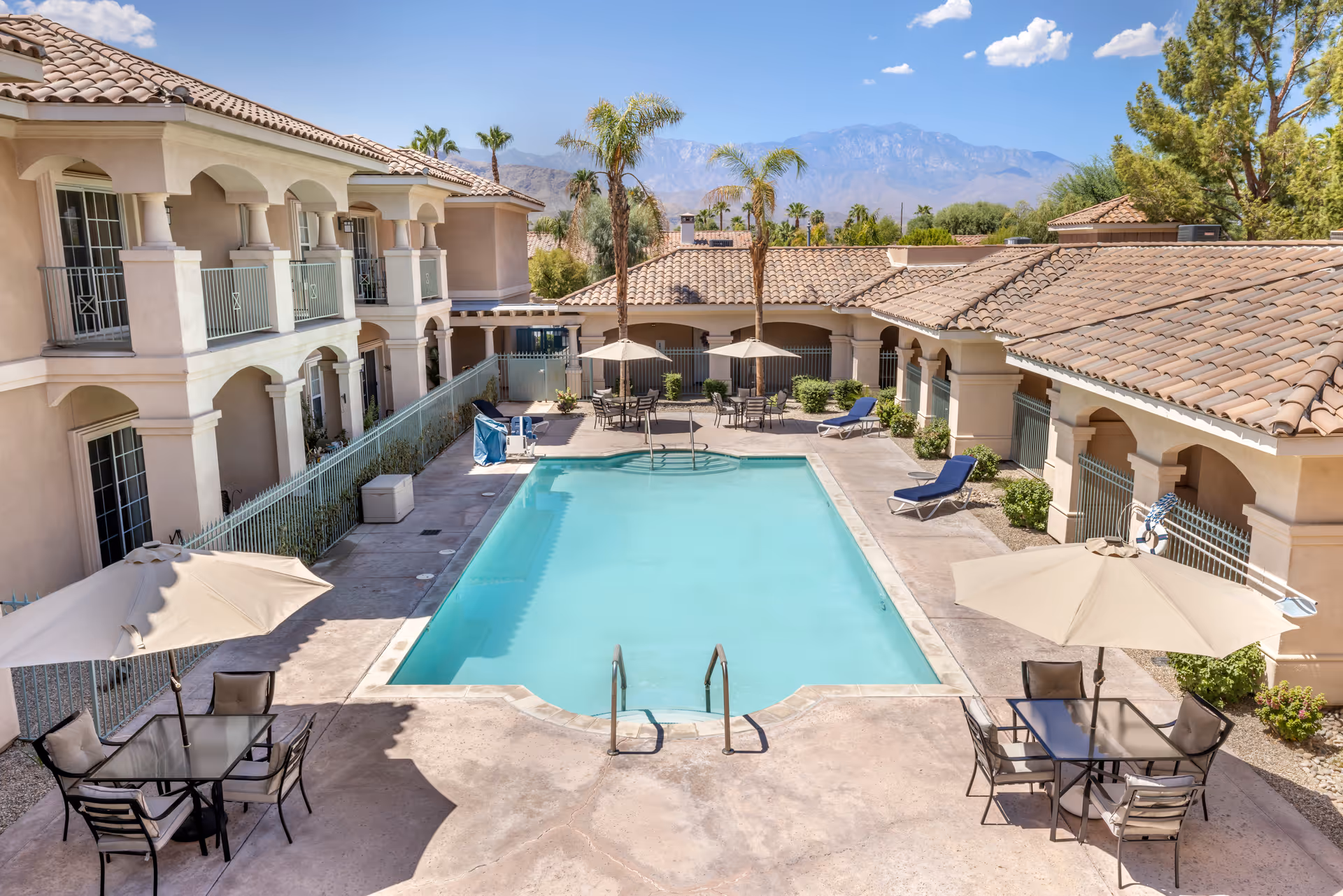 Outdoor swimming pool area at Brookdale Mirage Inn with lounge chairs, tables with umbrellas, surrounded by a two-story building with arches and a tiled roof, palm trees, and mountains in the background under a clear blue sky.