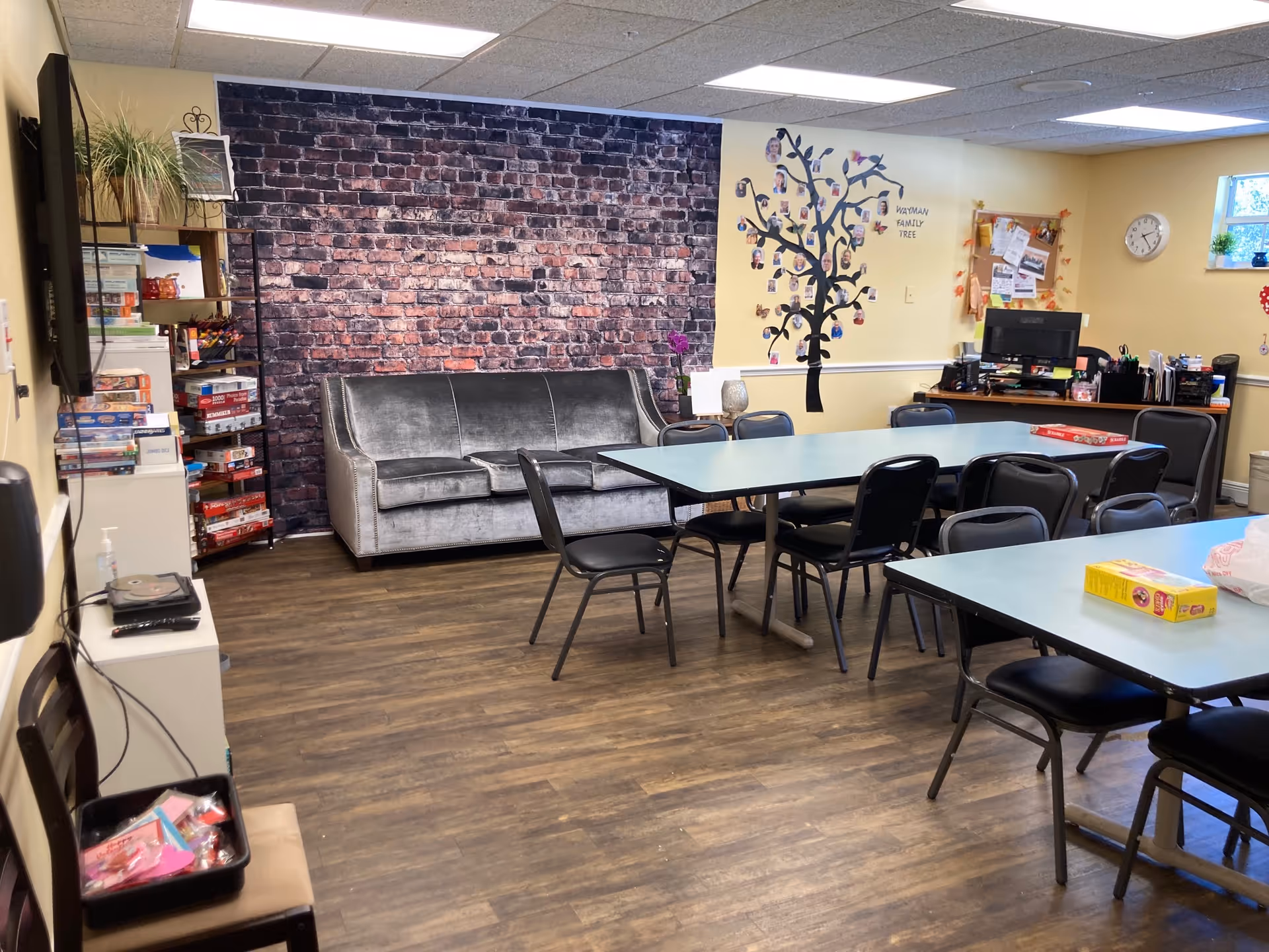 A common room in a senior living facility with a gray couch against a brick-patterned wall, several black chairs around two light blue tables, a shelving unit with board games, a desk with a computer, and a wall decorated with a family tree labeled 'Wayman Family Tree'.