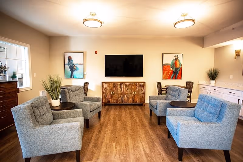 Bright communal lounge with four upholstered armchairs and side tables facing a wall-mounted TV above a decorative cabinet.
