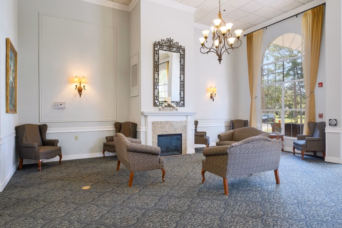 A bright and elegant senior living facility common area with patterned carpet, several upholstered armchairs arranged around a fireplace with a decorative mirror above it, wall sconces, a chandelier, and large arched windows with beige curtains letting in natural light.