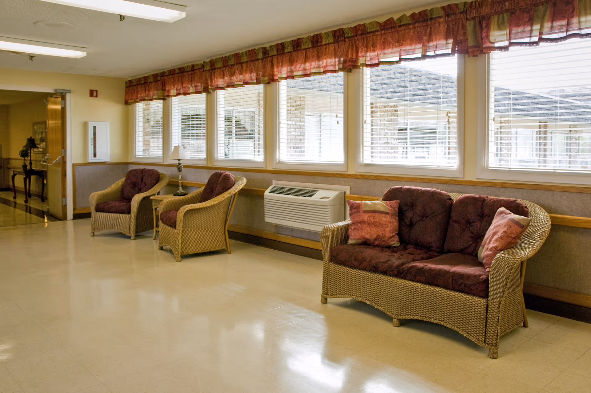 A hallway seating area in a senior living facility with wicker furniture including a loveseat and two armchairs with maroon cushions and decorative pillows. There is a small table with a lamp between the armchairs. Large windows with blinds and a valance let in natural light, and a wall-mounted air conditioning unit is visible below the windows.