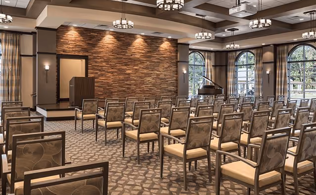 A large meeting or event room with rows of chairs facing a small stage with a podium. The room features a stone accent wall behind the podium, multiple ceiling lights, large arched windows with curtains, and a grand piano near the windows.