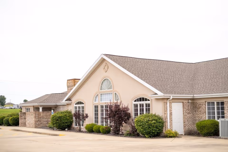 Exterior view of a single-story building with beige walls and a brown shingled roof. The building features large arched windows with white frames and is surrounded by neatly trimmed bushes and small trees. The foreground shows a paved parking area.