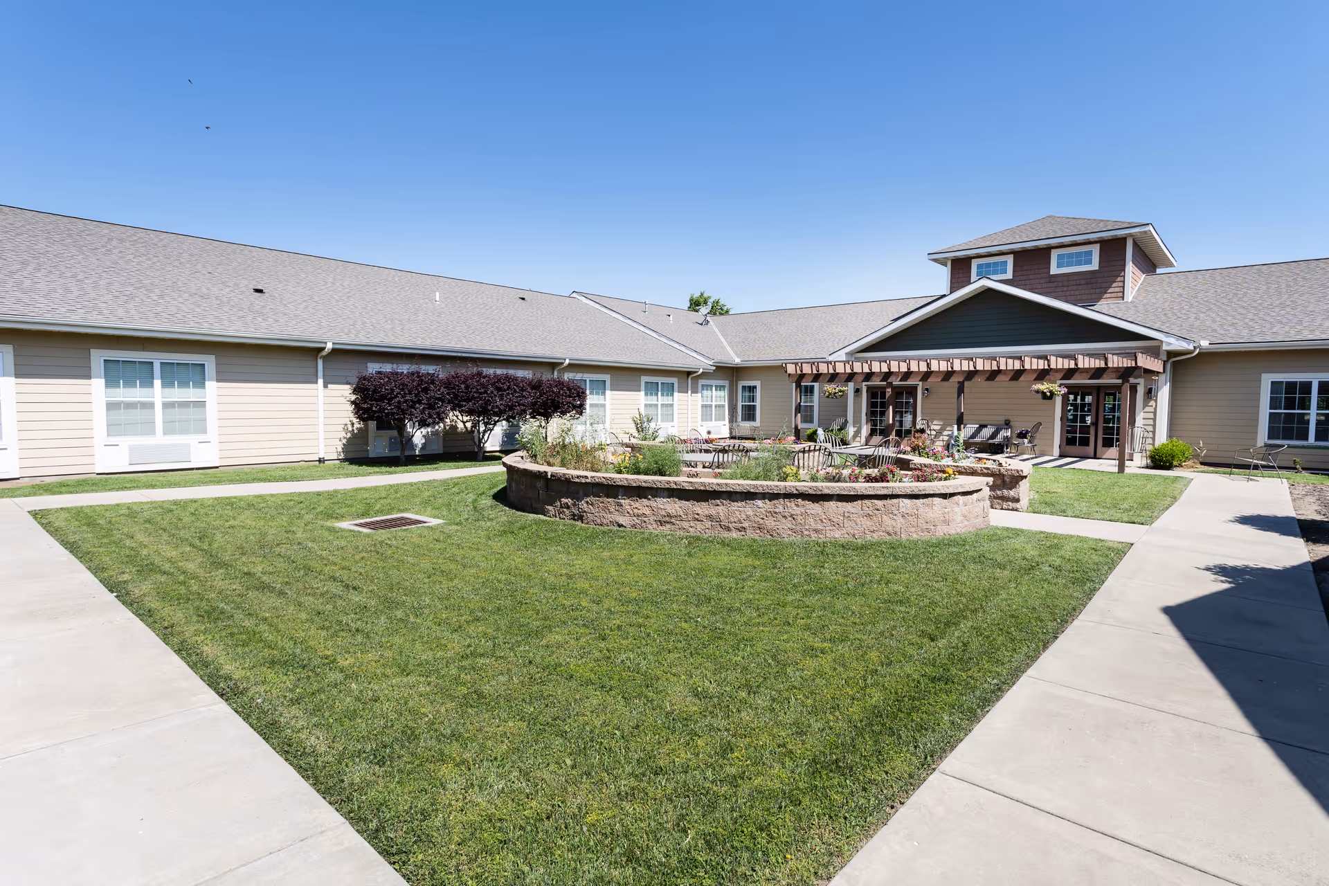 Outdoor courtyard area of Oak Pointe of Carthage with a well-maintained green lawn, a circular raised garden bed with plants, surrounded by paved walkways, and a building with beige siding and multiple windows under a clear blue sky.