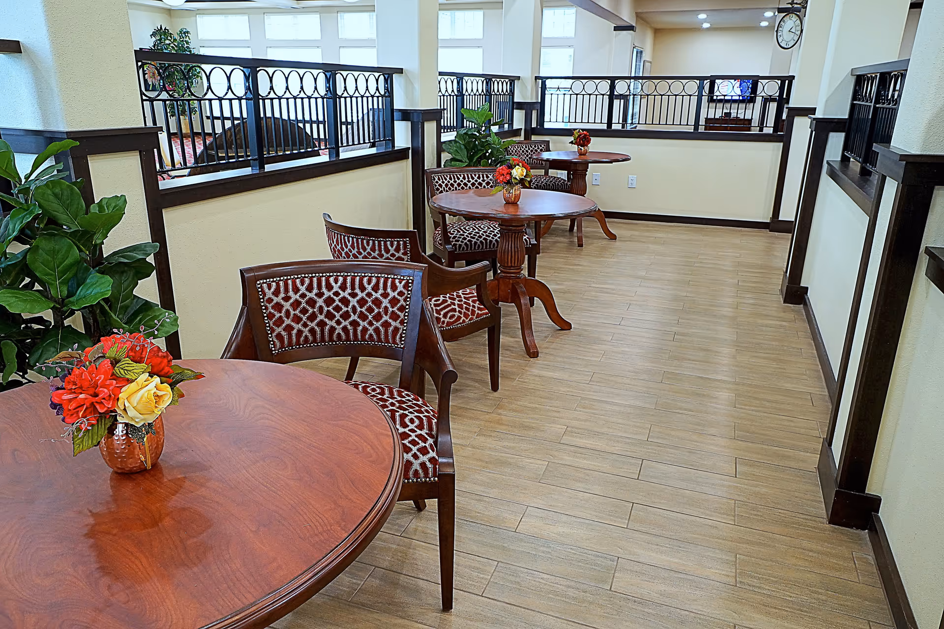 A hallway area in a senior living facility with round wooden tables and patterned chairs. Each table has a small floral arrangement. The floor is wood-patterned tile, and there are railings along the half walls separating this area from an adjacent room with windows and a TV.