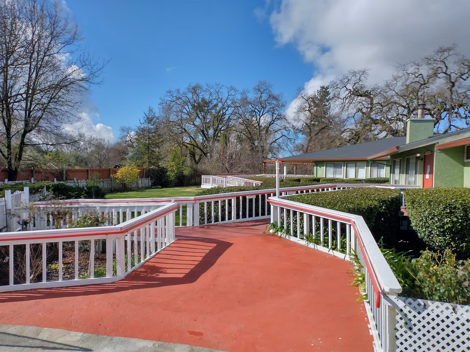 Outdoor view of a senior living facility with a red paved walkway and white railings leading to a green building. The area is surrounded by bushes, trees, and a clear blue sky with some clouds.
