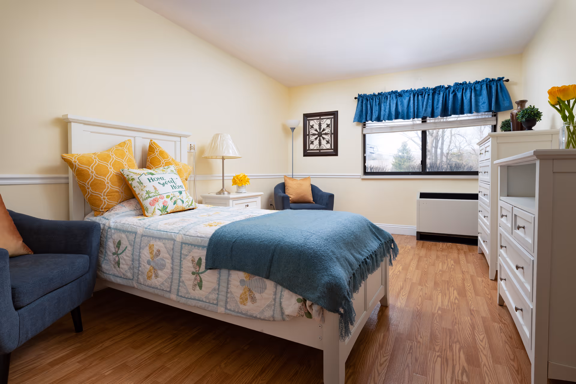 A cozy bedroom with a single white bed adorned with a floral quilt, yellow and patterned pillows, and a teal throw blanket. The room features light yellow walls, a wooden floor, a blue armchair with an orange pillow, a white nightstand with a lamp and yellow flowers, a white dresser, and a window with blue curtains letting in natural light.