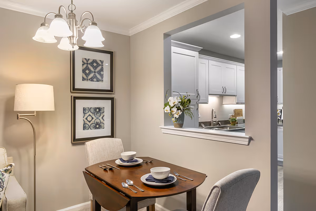 Small dining area set for two with a wooden table, upholstered chairs, framed wall art, a floor lamp, and a pass-through to a white kitchen.