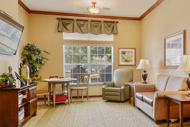 Cozy senior living common room with a sofa, recliner, small table and chairs, TV cabinet, and a large window.