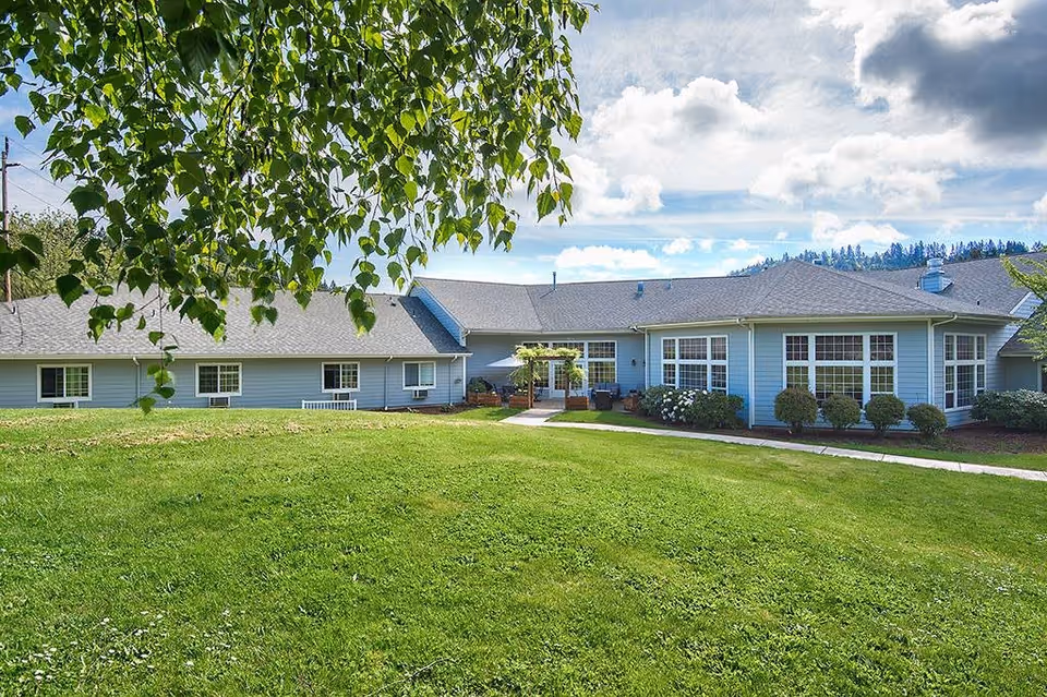 Exterior view of a single-story assisted living facility building with light blue siding, multiple large windows, and a well-maintained green lawn in the foreground. A tree branch with green leaves partially frames the top left corner of the image under a partly cloudy sky.