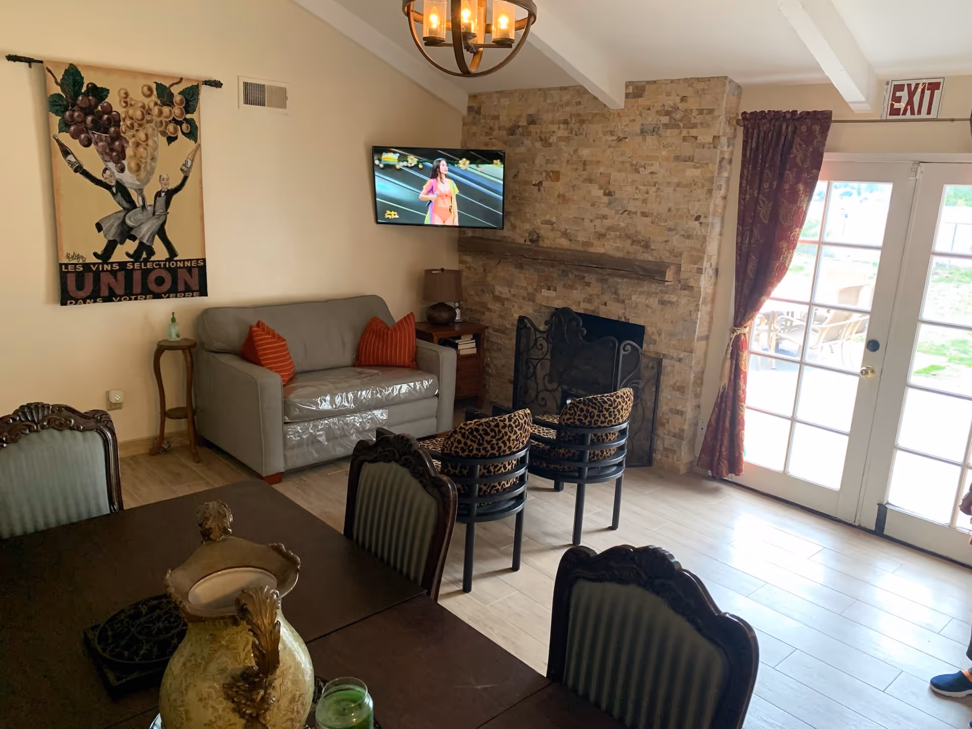 A cozy living room with a stone fireplace, two leopard print chairs, a gray loveseat with two orange pillows, a wall-mounted TV, and a dining table with ornate chairs. There is a decorative wall hanging with grapes and French text, a chandelier, and glass doors with red curtains leading outside.