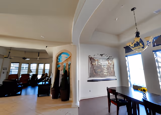 Interior view of a senior living facility showing a dining area with a dark wooden table and chairs near large windows with blinds. A decorative light fixture hangs above the table. Adjacent to the dining area is a lounge space with multiple chairs and large windows allowing natural light to fill the room. The walls are painted white and there is a decorative wall hanging visible.