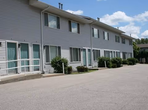 Two-story light gray building exterior with teal doors, railings, and shrubs.