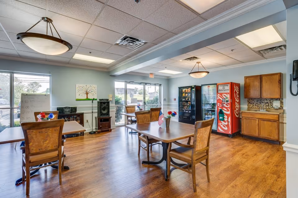 A bright dining area in a senior living facility with wooden floors and light blue walls. The room contains several wooden tables and chairs, a Coca-Cola vending machine, a snack vending machine, a small cabinet with a sink, and large windows letting in natural light. There are ceiling lights and a small American flag on one of the tables.
