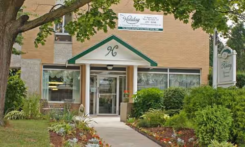Exterior view of a senior living facility entrance with green shrubs and landscaping.
