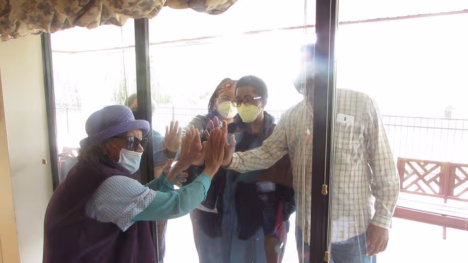 An elderly woman wearing a purple hat, sunglasses, and a face mask inside a building presses her hands against a glass door, touching hands with three people wearing face masks on the outside. The people outside are standing close to the glass door, engaging in a moment of connection.
