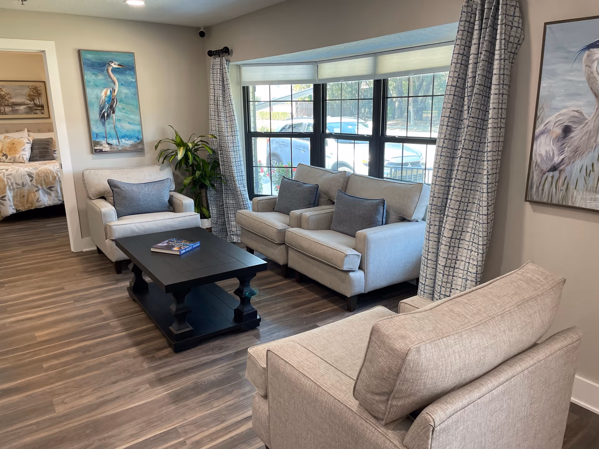 A cozy living room with three beige armchairs and a matching loveseat arranged around a black wooden coffee table with a book on it. Large windows with patterned curtains let in natural light. A green plant is placed near the corner, and two paintings of birds hang on the walls. A bedroom with a bed and floral bedding is visible through an open doorway.