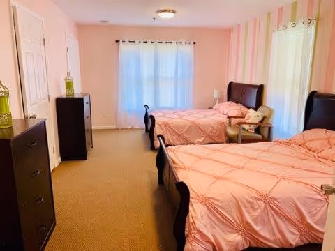 A bedroom with two twin beds covered in pink comforters, a beige armchair with pillows between the beds, a window with sheer white curtains, a striped pink and white accent wall, and two dark wood dressers on the left side of the room.