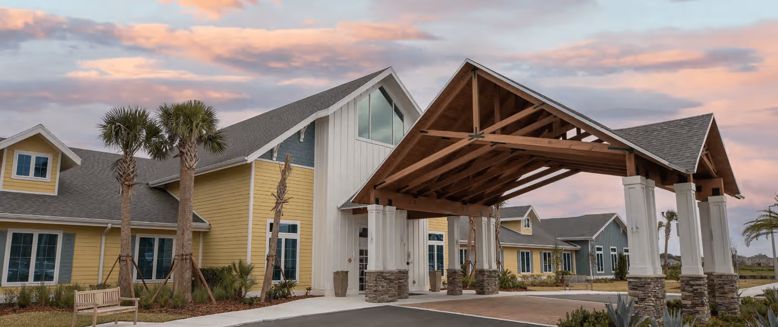 Exterior view of a senior living facility building with a large covered entrance supported by white columns with stone bases. The building has yellow and blue siding with multiple windows and a gray shingled roof. There are palm trees and landscaping around the entrance, and a wooden bench is visible on the left side. The sky is partly cloudy with a warm sunset glow.