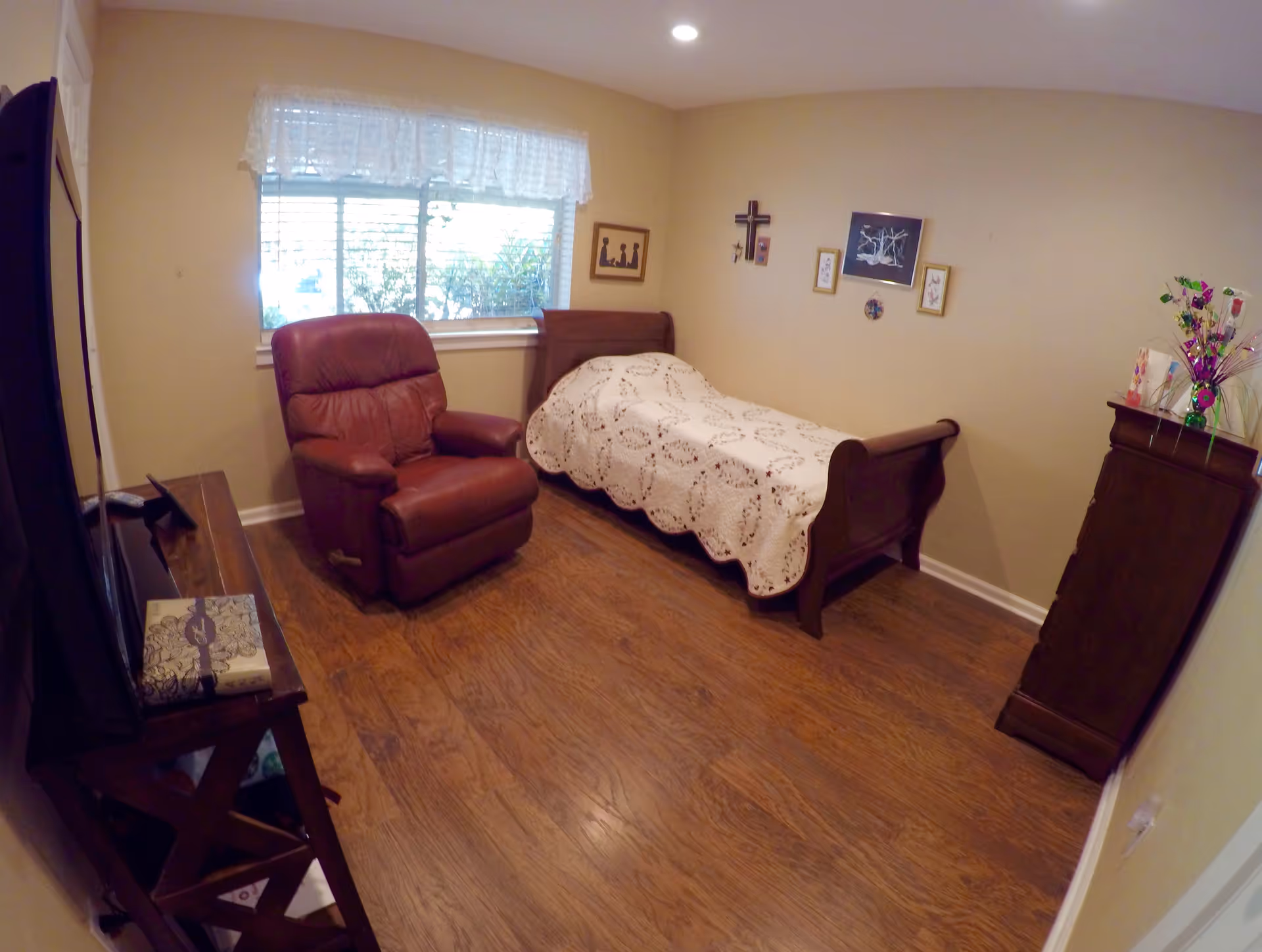 A small bedroom with wooden flooring featuring a single bed with a white crocheted blanket, a brown recliner chair, a wooden dresser with decorative items, and a window with white lace curtains. The walls have framed pictures and a cross hanging.