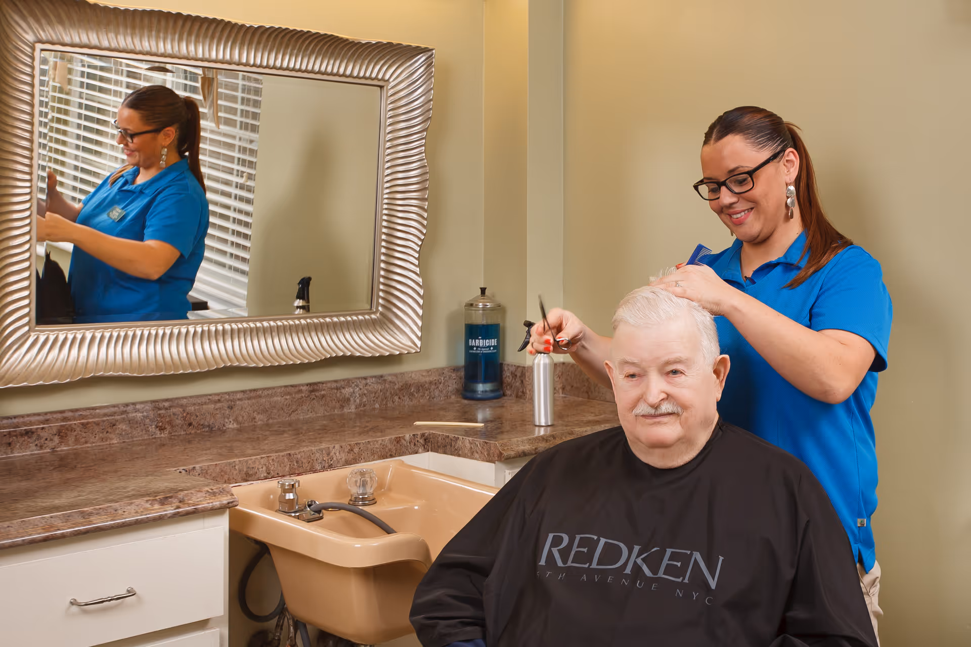 A hairstylist wearing a blue shirt is cutting the hair of an elderly man seated in a chair with a black Redken cape. They are in a room with a beige wall, a large decorative mirror, and a countertop with a sink and hair care products.