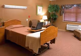 A furnished bedroom with a wooden bed, laptop on desk, chair, lamp, and window blinds.