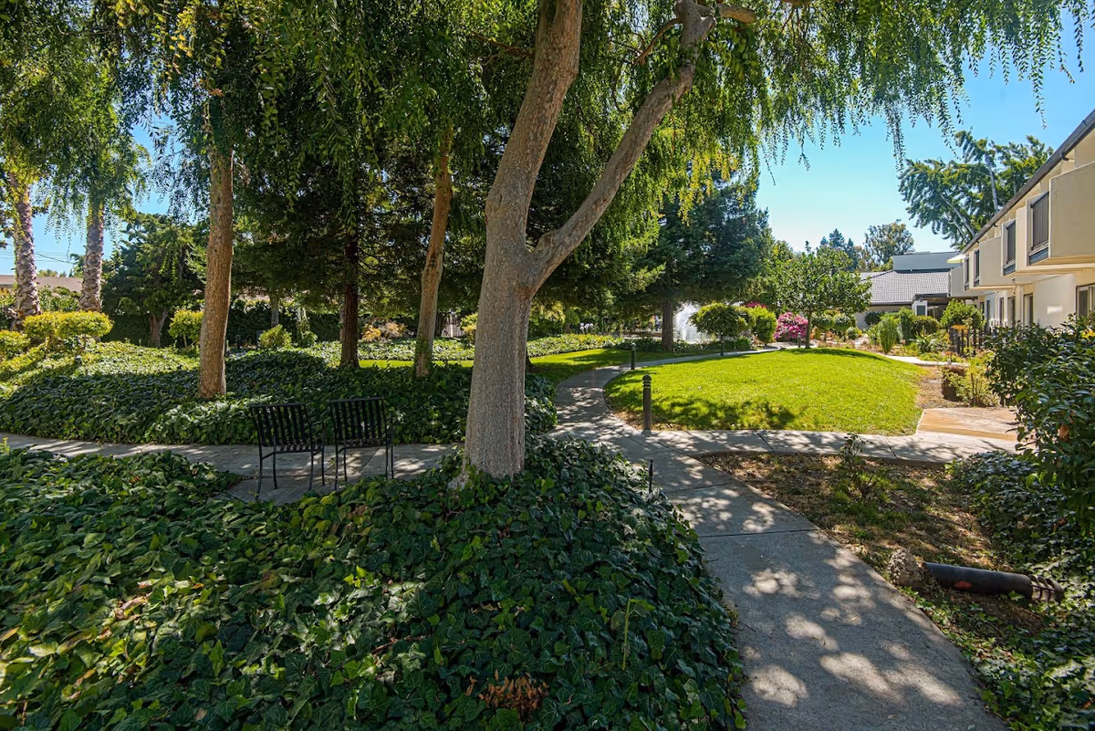 A sunny outdoor garden area at Villa Serena Retirement Community featuring a winding concrete pathway, lush green grass, various trees and shrubs, and two black metal chairs placed on the path. Residential buildings are visible on the right side under a clear blue sky.