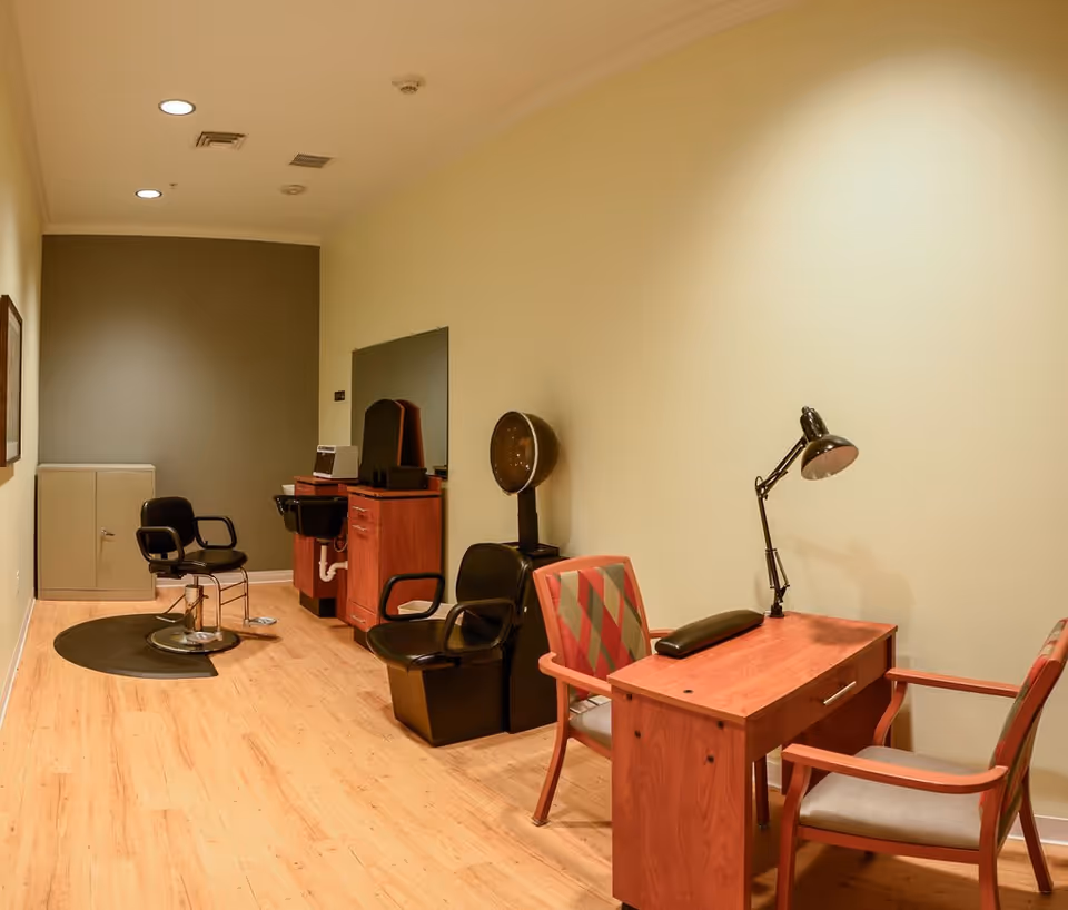 Interior view of a salon area in a senior living facility with wooden flooring, two black salon chairs, a hair washing station, a hair dryer chair, and a wooden desk with two chairs and a desk lamp.