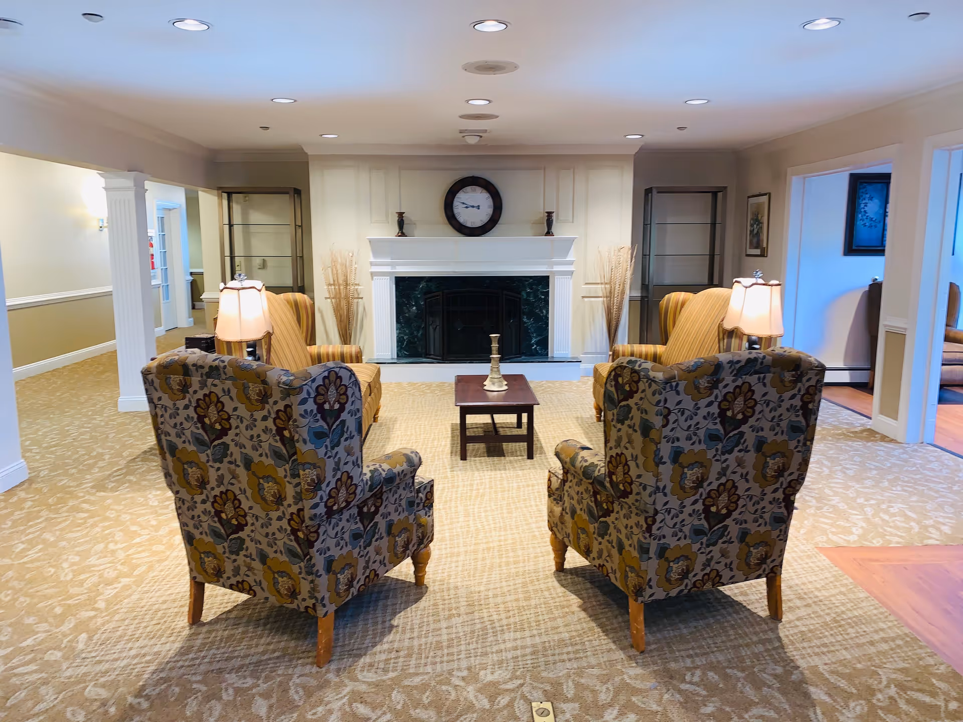 A cozy sitting area with four upholstered armchairs arranged around a small wooden coffee table. The chairs have floral and striped patterns. Behind the seating area is a white fireplace with a dark green marble surround, flanked by two glass display cabinets. A round wall clock and decorative candle holders are on the mantel. The room has beige patterned carpet and soft lighting from ceiling lights and table lamps.