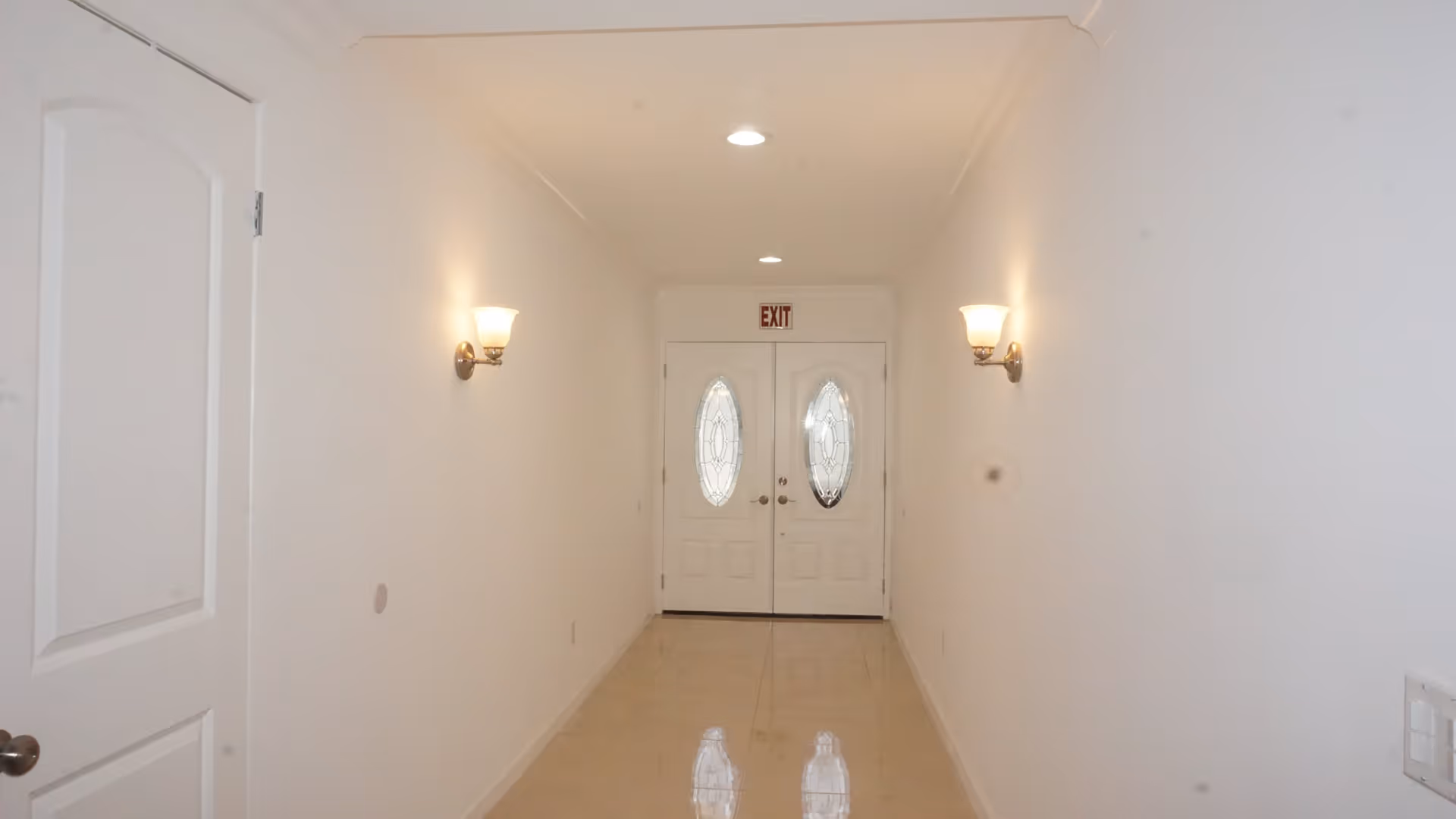 A narrow, well-lit hallway with white walls and a shiny tiled floor leading to a set of double doors with oval glass panels. Two wall-mounted light fixtures are on either side of the hallway, and an exit sign is above the doors.