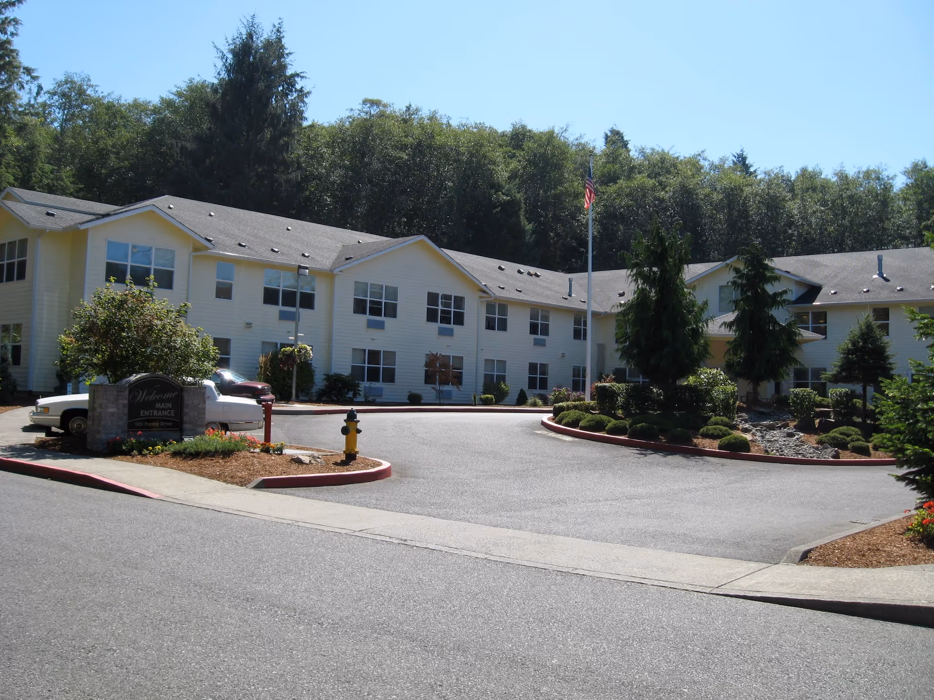 Front exterior of a two-story assisted living building with a circular driveway, landscaped island, and an American flag.