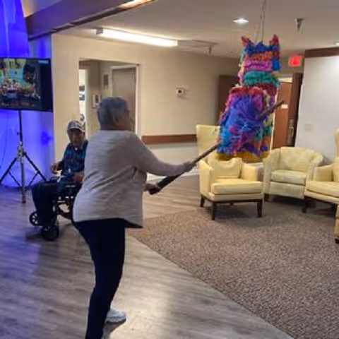 A woman swings a stick at a colorful piñata in a senior living facility common room while others watch.