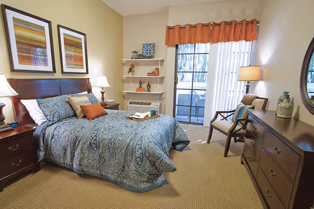A cozy bedroom with a large bed covered in a blue patterned comforter and multiple pillows. The room features two framed artworks above the headboard, two bedside tables with lamps, a small shelving unit with decorative items, a window with sheer curtains and an orange valance, a wooden chair with a cushion, and a wooden dresser with decorative vases and a round mirror above it.