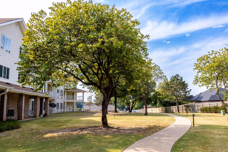A curved concrete walkway runs through a grassy outdoor area with several trees. On the left side, there is a multi-story residential building with balconies and brick pillars. The sky is partly cloudy with blue and white hues.