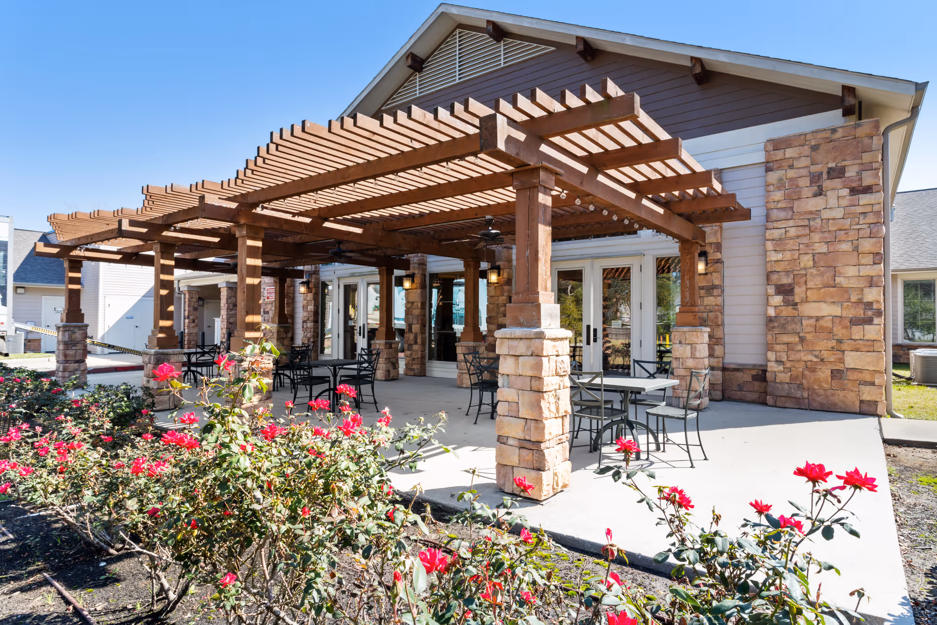 Outdoor patio with a wooden pergola, tables and chairs in front of a stone-faced building and red flowering bushes.