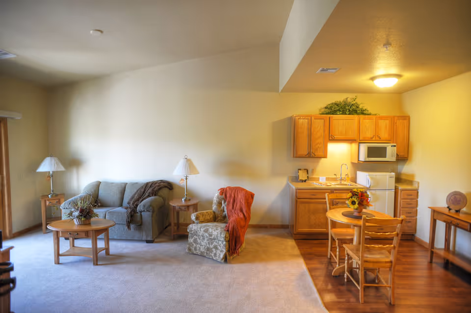 A furnished living area with a sofa, armchair, coffee table and lamps next to a small kitchenette and dining table.