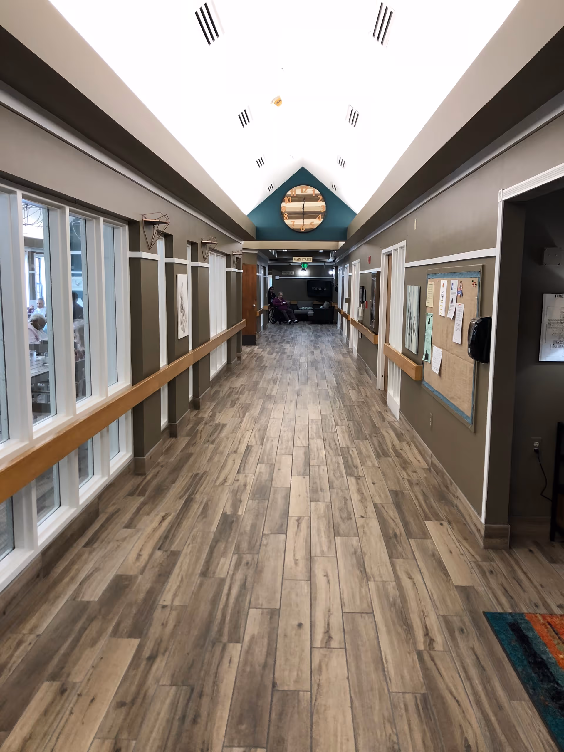 A long, well-lit hallway with wood-patterned flooring and beige walls featuring white trim. On the left side, there are large windows looking into a dining area where people are seated. On the right side, there is a bulletin board with various papers pinned to it. At the end of the hallway, a large round clock is mounted on a teal accent wall above a sitting area with a person in a wheelchair and another person nearby.